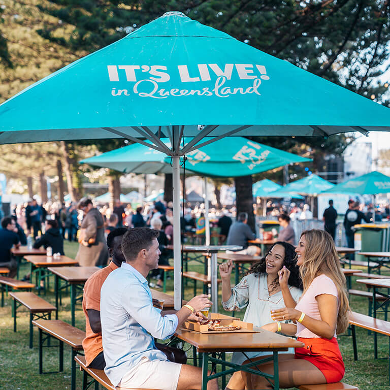 Friends enjoying food and drinks at an outdoor event in Queensland under a turquoise �It's Live! in Queensland� umbrella.